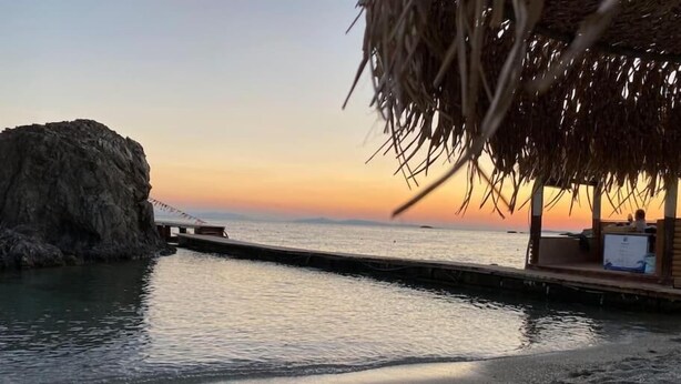 Una playa con un promontorio rocoso, un muelle de madera y una estructura de techo de paja.