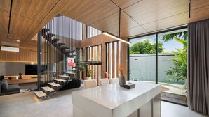 A modern interior with a central island, a staircase, and large windows offering a view of greenery. - Alex Villas Complex N2 (Canggu)