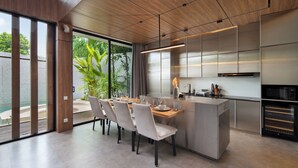 A modern kitchen with a central island, large windows, and a dining area with chairs. - Alex Villas Complex N2 (Canggu)