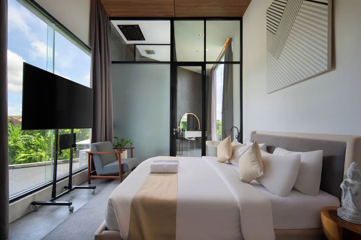 A modern hotel room with a large bed, a flat-screen TV, a seating area with a table and chairs, and a view of greenery outside.