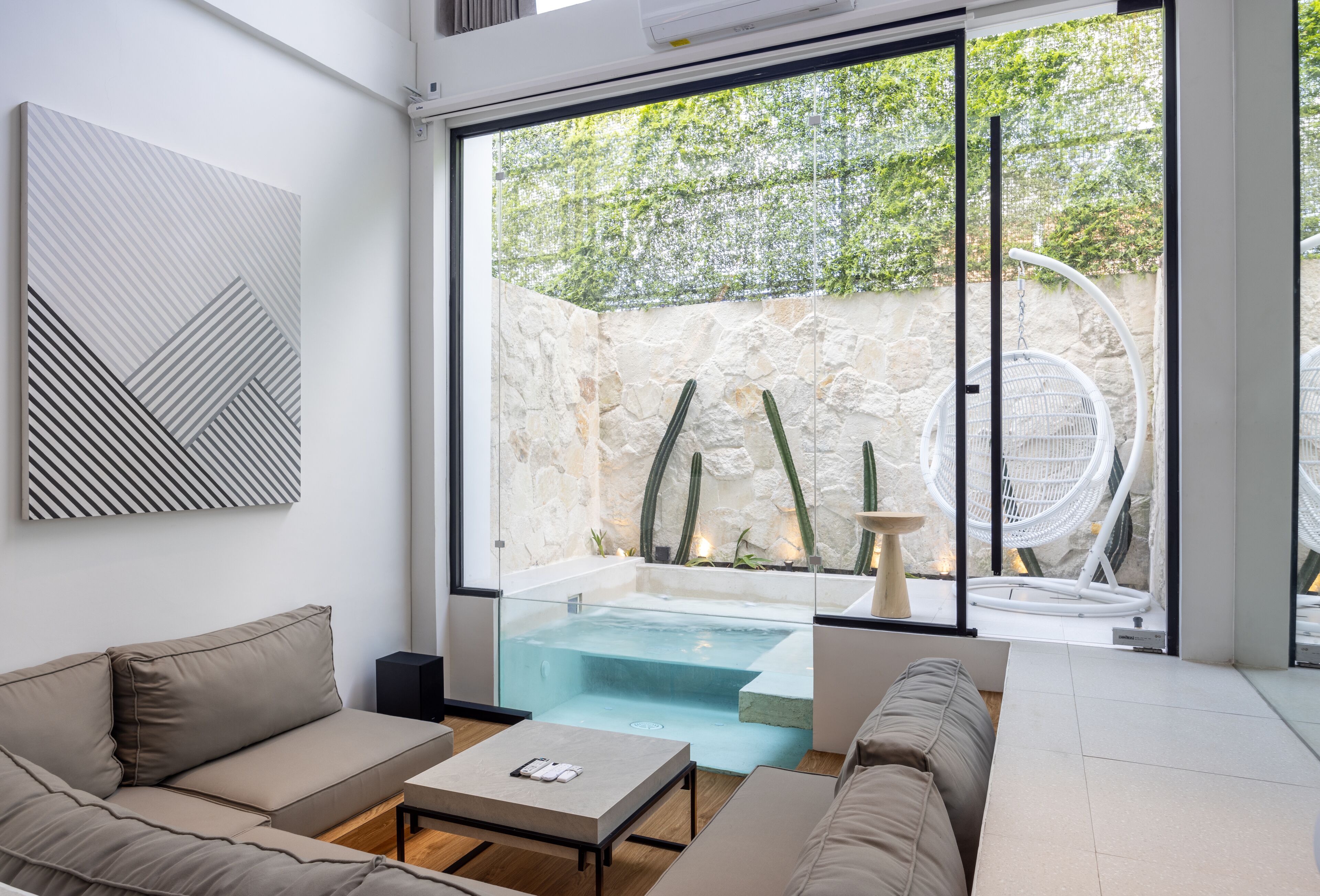 A modern living room with a glass wall, a beige sofa, a coffee table, and a stone wall with a cactus garden visible through the glass.