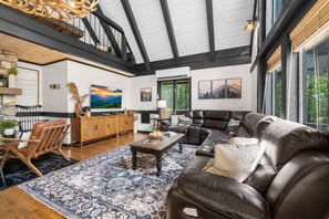 A spacious living room with a high ceiling, a leather sofa, a wooden coffee table, and a large window providing a view of the outdoors.