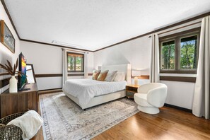 A modern bedroom with a large bed, bedside tables, a chair, and a view of greenery through the windows.