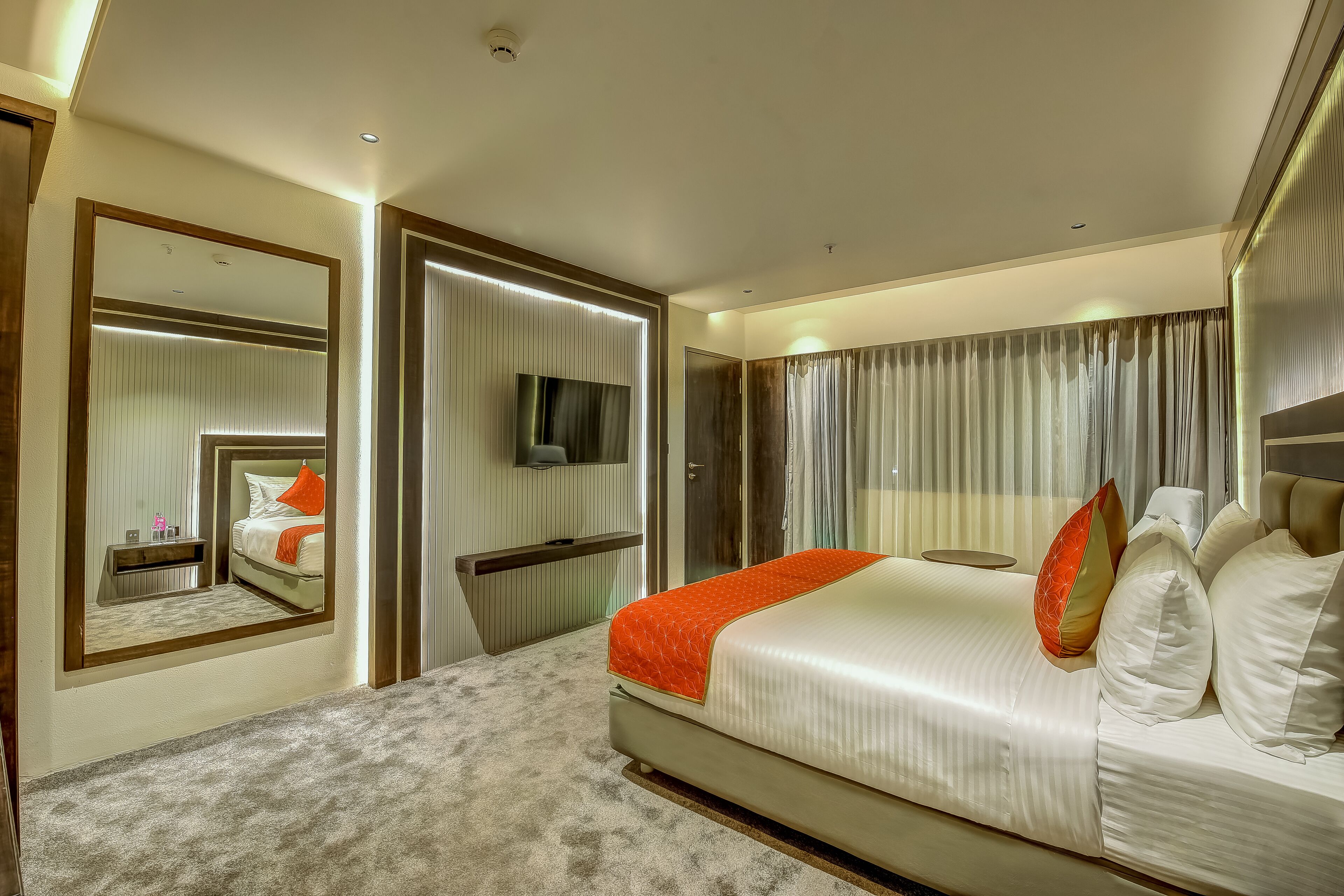 A modern hotel room with a large bed, a TV, a mirror, and a window with curtains.