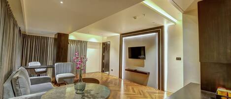 A modern living room with a round table, sofa, and chairs. There is a TV mounted on the wall and a wooden floor.