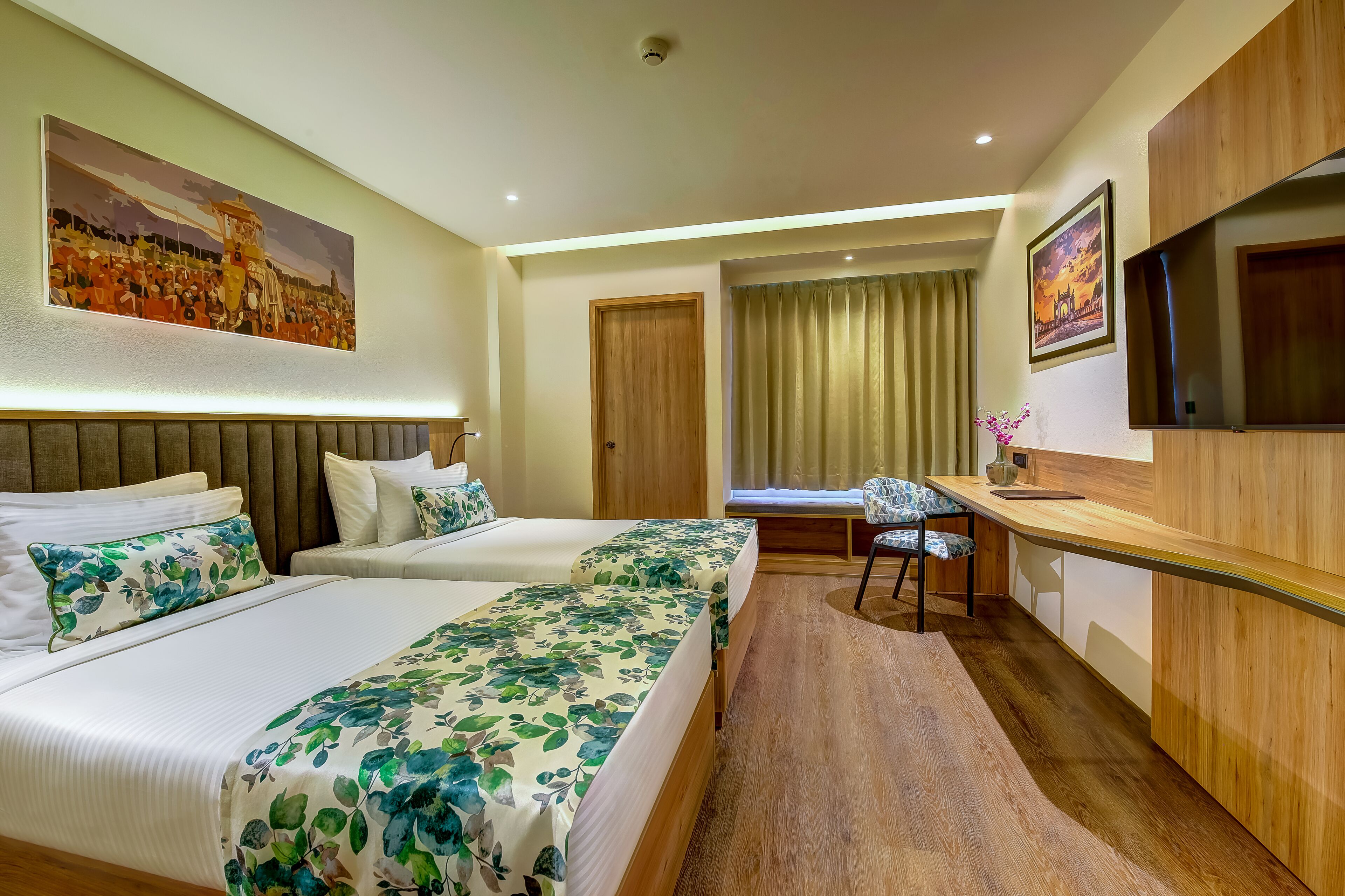 A hotel room with two beds, a desk, a chair, a TV, and artwork on the walls.