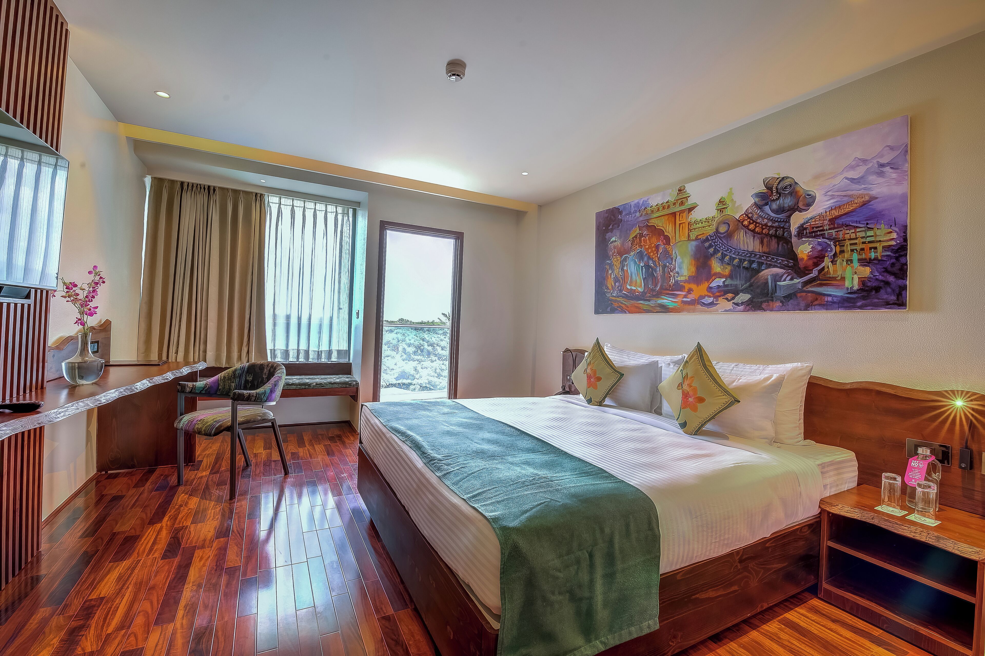 Habitación de hotel con piso de madera, cama con manta verde, escritorio con silla, un cuadro en la pared y ventana con cortinas.