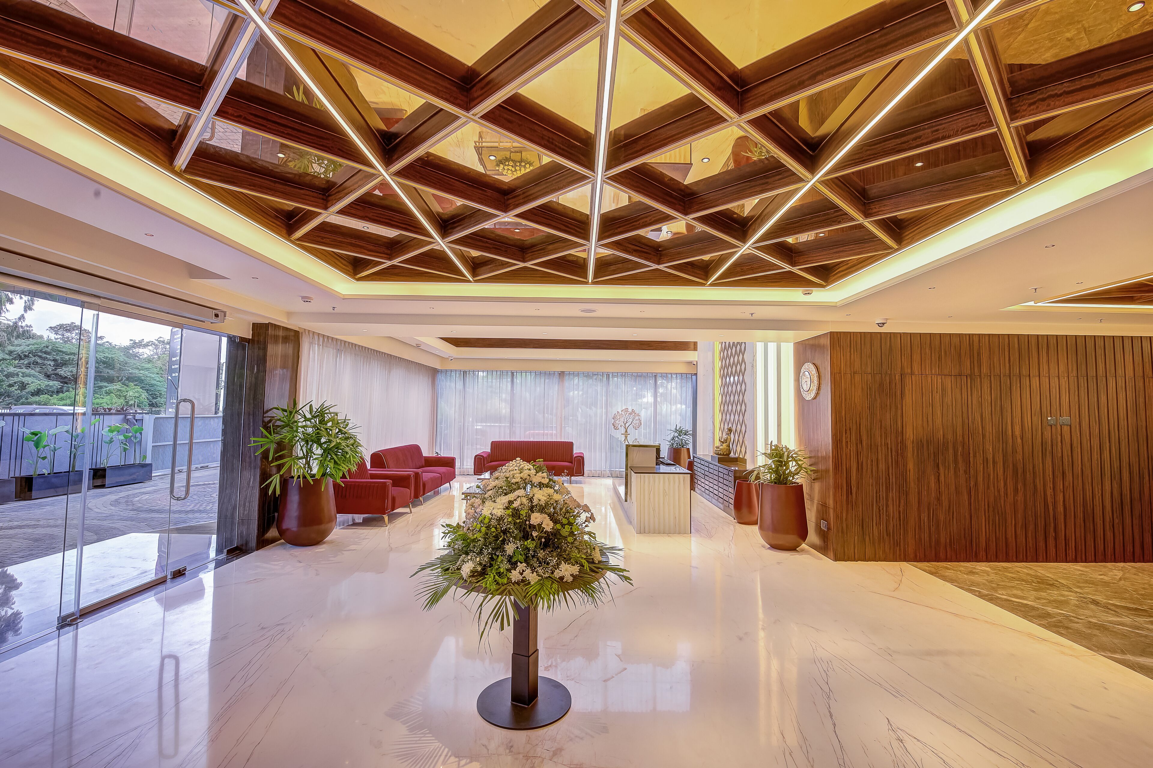 Een moderne lobby met een geometrisch plafondontwerp, een receptiebalie, rode zitplaatsen en potplanten.
