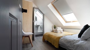A bedroom with a skylight, a bed with a yellow blanket, a white chair, and a bathroom visible through an open door. - Get Away Home With Hot Tub Sleeps 7 (Peacehaven)