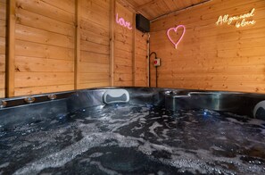 A wooden cabin with a hot tub, neon signs reading "love" and "All you need is love," and a heart-shaped neon light.