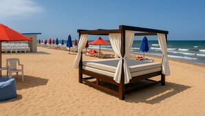 Un lit installé sur la plage, surmonté d’un dais, entouré de chaises de plage et de parasols, avec l’océan en arrière-plan.