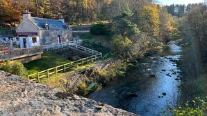 La Casa Fiddichside, un edifici de pedra amb façana blanca i porta vermella, situada al costat d'un riu amb un pont de fusta que hi通.