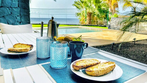 Une table dressée pour le petit-déjeuner, avec des toasts, une cafetière et un saladier de fruits, placée près d’une piscine, offrant une vue sur des palmiers et une clôture.