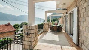 Ein Balkon mit Blick auf das Meer und die Berge, Steinmauern, ein gefliester Boden und ein Tisch mit Stühlen.