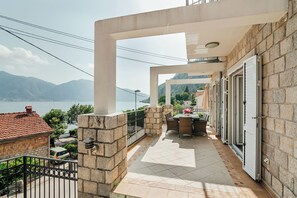 Uma varanda com vista para o mar e montanhas, paredes de pedra, piso de azulejos e uma mesa com cadeiras.