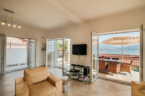 Uma sala de estar com sofá, mesa de centro de vidro, televisão de tela plana e varanda com mobiliário externo.