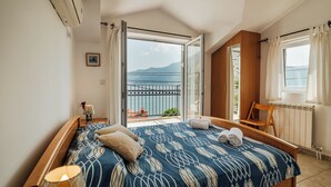 Ein Schlafzimmer mit Bett, Nachttisch, Lampe, Fenster mit Vorhängen und Blick auf einen Balkon.
