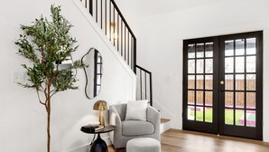 A modern living room with a staircase, a grey armchair, a round ottoman, and a potted plant.