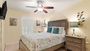Une chambre à coucher comprenant un lit, une table de chevet, une lampe, un ventilateur de plafond et des œuvres d’art accrochées aux murs.