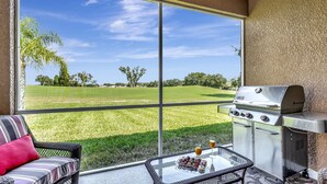 Ein Balkon mit Grill, einem Glastisch mit Speisen und Getränken sowie Blick auf ein grünes Feld.