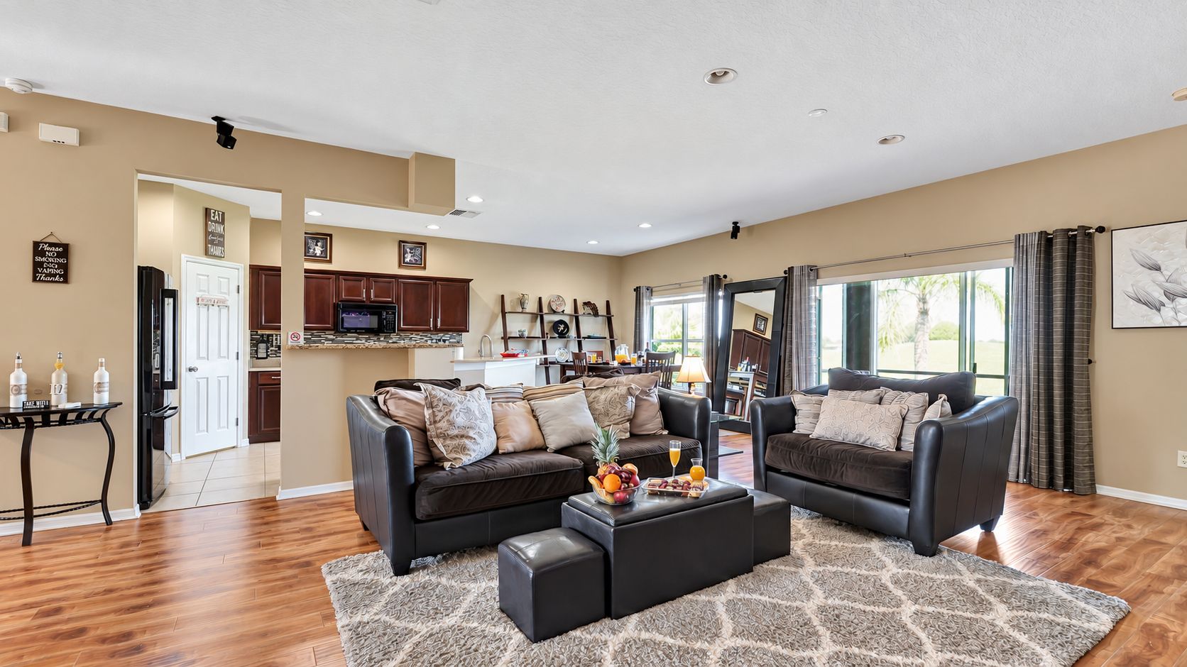 A spacious living room with a dark brown leather sofa set, a coffee table with fruit and drinks, and a kitchen in the background.