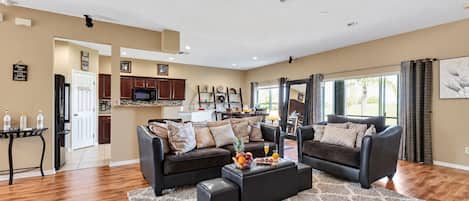 A spacious living room with a dark brown leather sofa set, a coffee table with fruit and drinks, and a kitchen in the background.