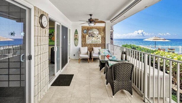 A balcony with a view of the ocean, a wicker chair, a table with a glass top, and a ceiling fan.