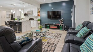 A modern living room with a black leather sofa, a glass coffee table, and a TV mounted on a dark blue wall.