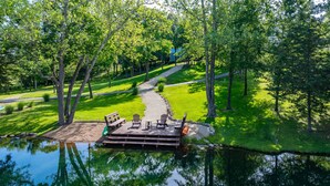 Um deck tranquilo à beira do lago, com um banco de madeira e cadeiras Adirondack, rodeado por vegetação exuberante e um caminho claro que leva a uma casa.