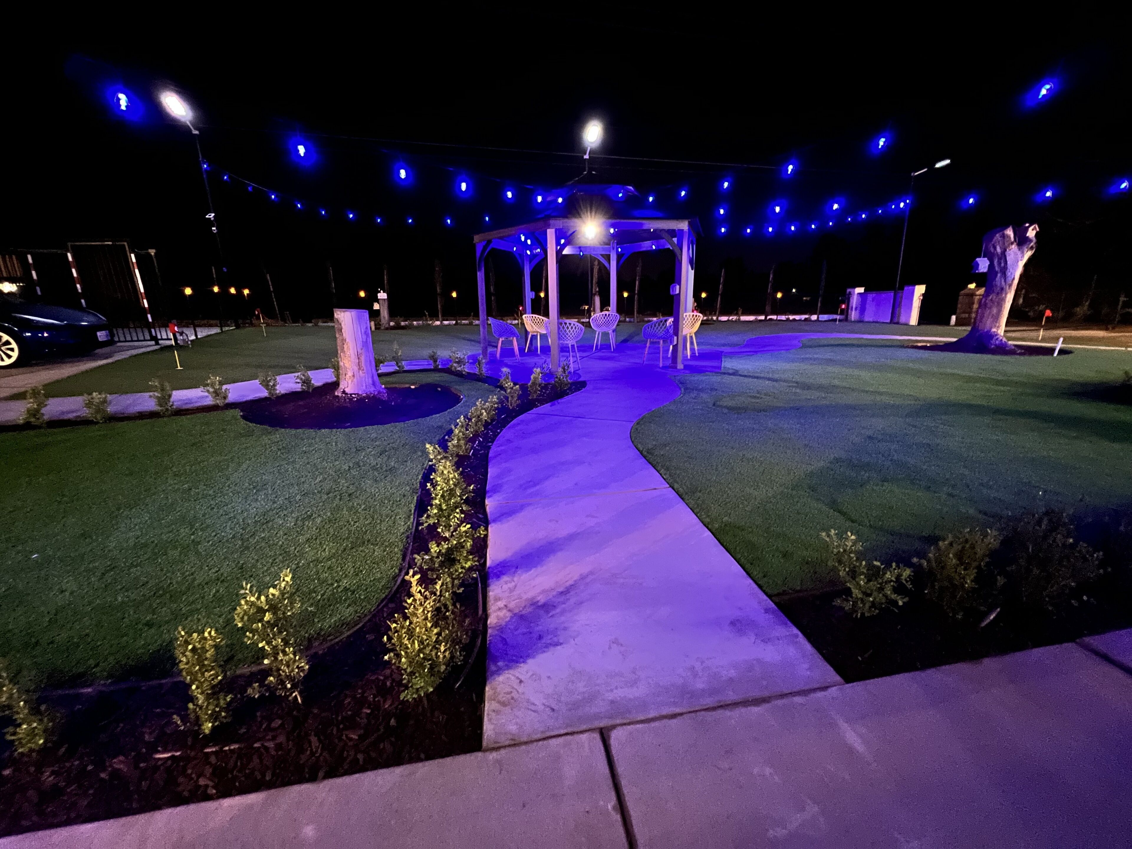 Un espacio al aire libre nocturno con un mirador, césped artificial y luces colgantes.