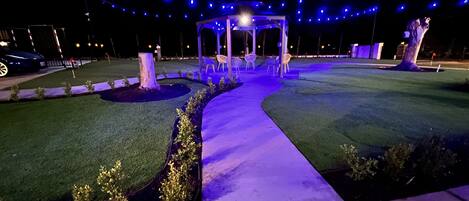 Un espacio al aire libre nocturno con un mirador, césped artificial y luces colgantes.