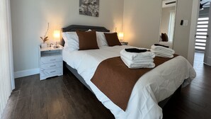 Una habitación de hotel con una cama grande, dos mesitas de noche, un tocador, un ventilador de techo, una obra de arte en la pared, un espejo y una puerta que da a otra habitación.
