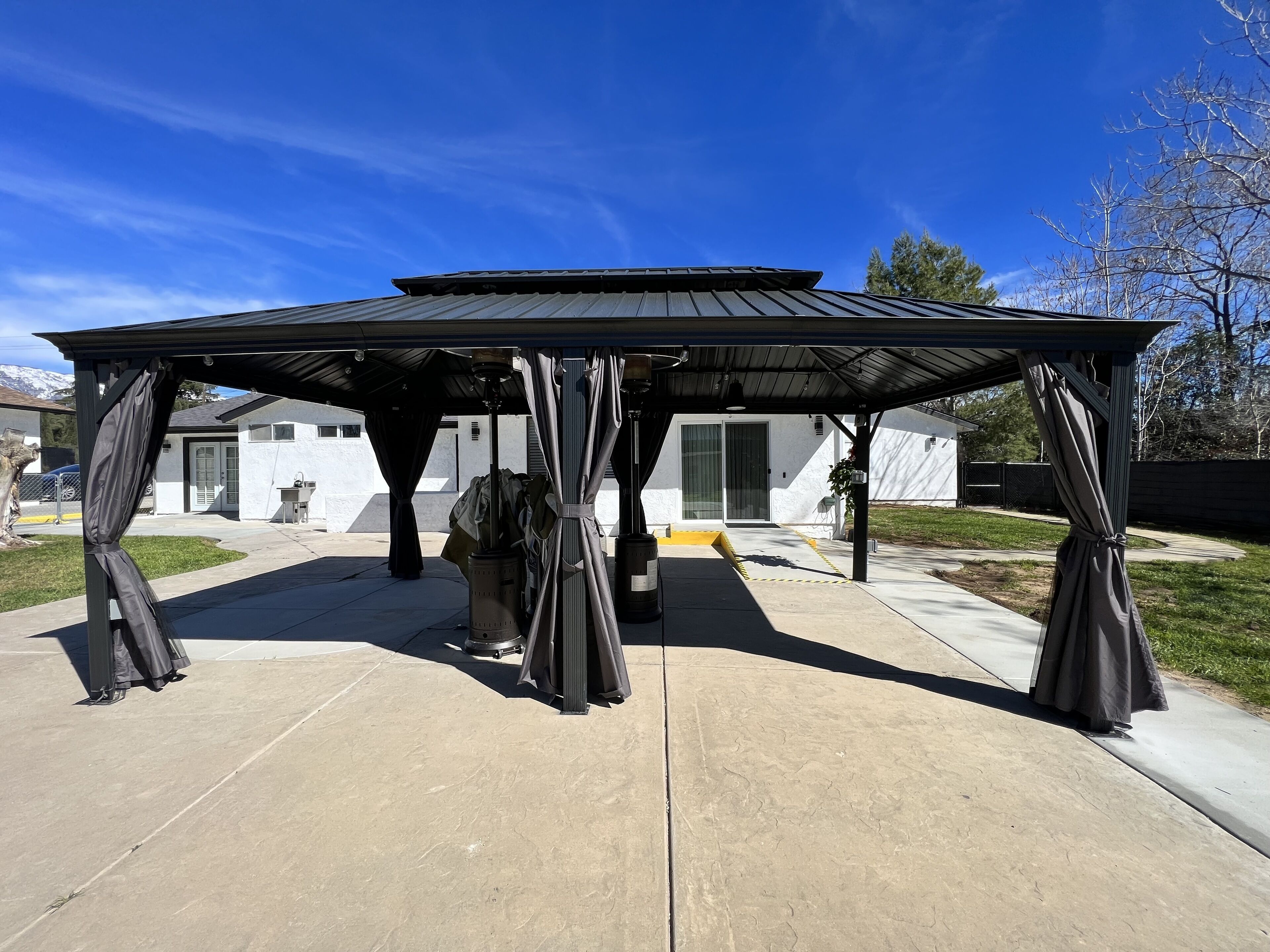Un patio cubierto con techo de metal y cortinas, suelo de concreto y vista a un edificio blanco al fondo.