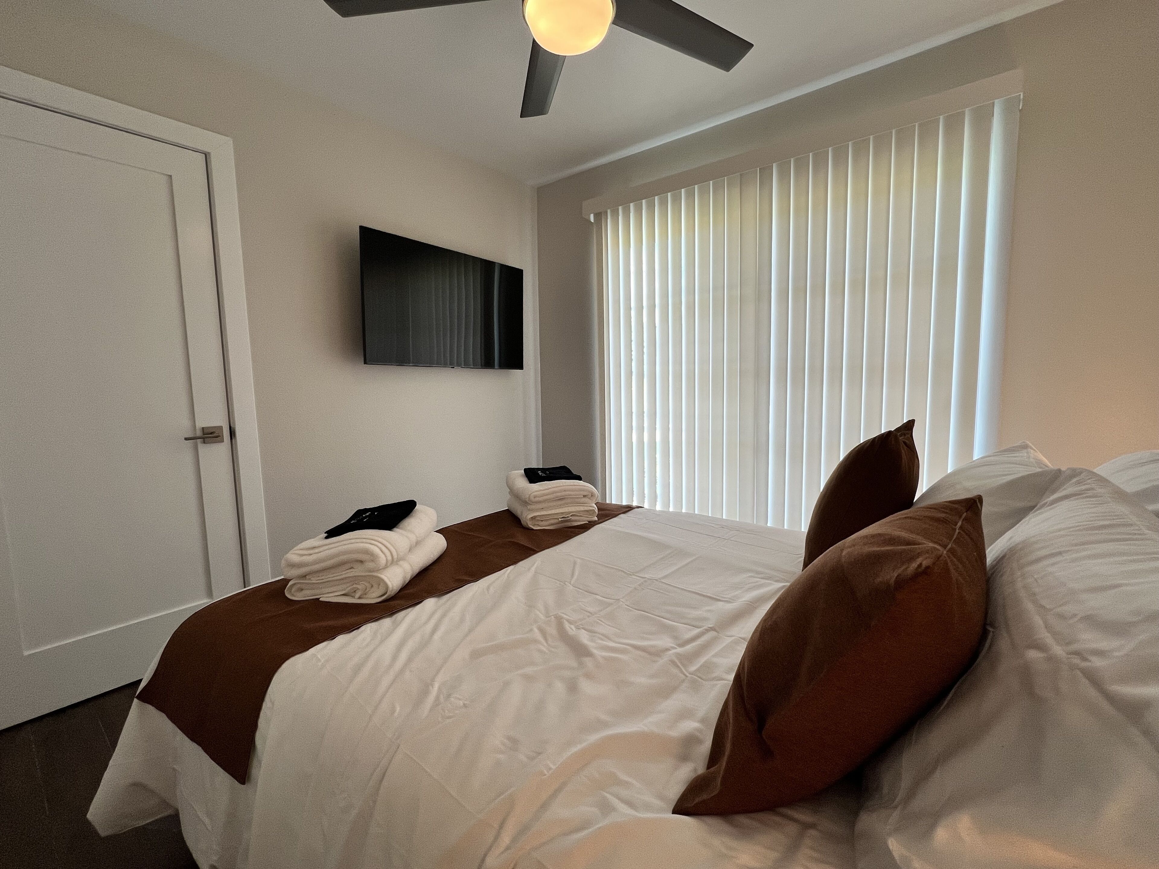 Una habitación de hotel con cama, televisor de pantalla plana montado en la pared, ventana con persianas y ventilador de techo.