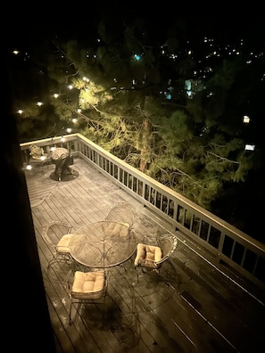 A wooden deck with outdoor furniture, including a round table and chairs, surrounded by trees and illuminated by string lights.