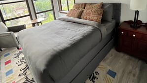 Un dormitorio con una cama grande, una mesita de noche, una lámpara y vistas a través de las ventanas.