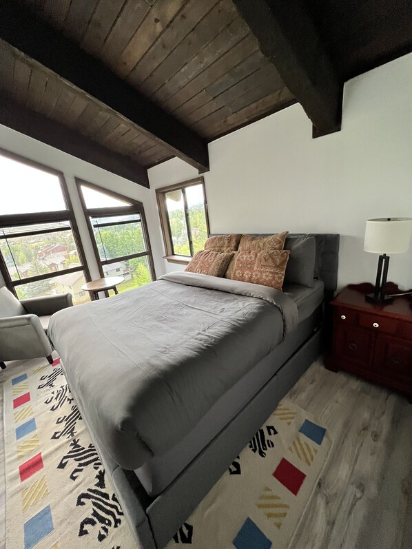 A bedroom with a large bed, a nightstand, a lamp, and a view through the windows.