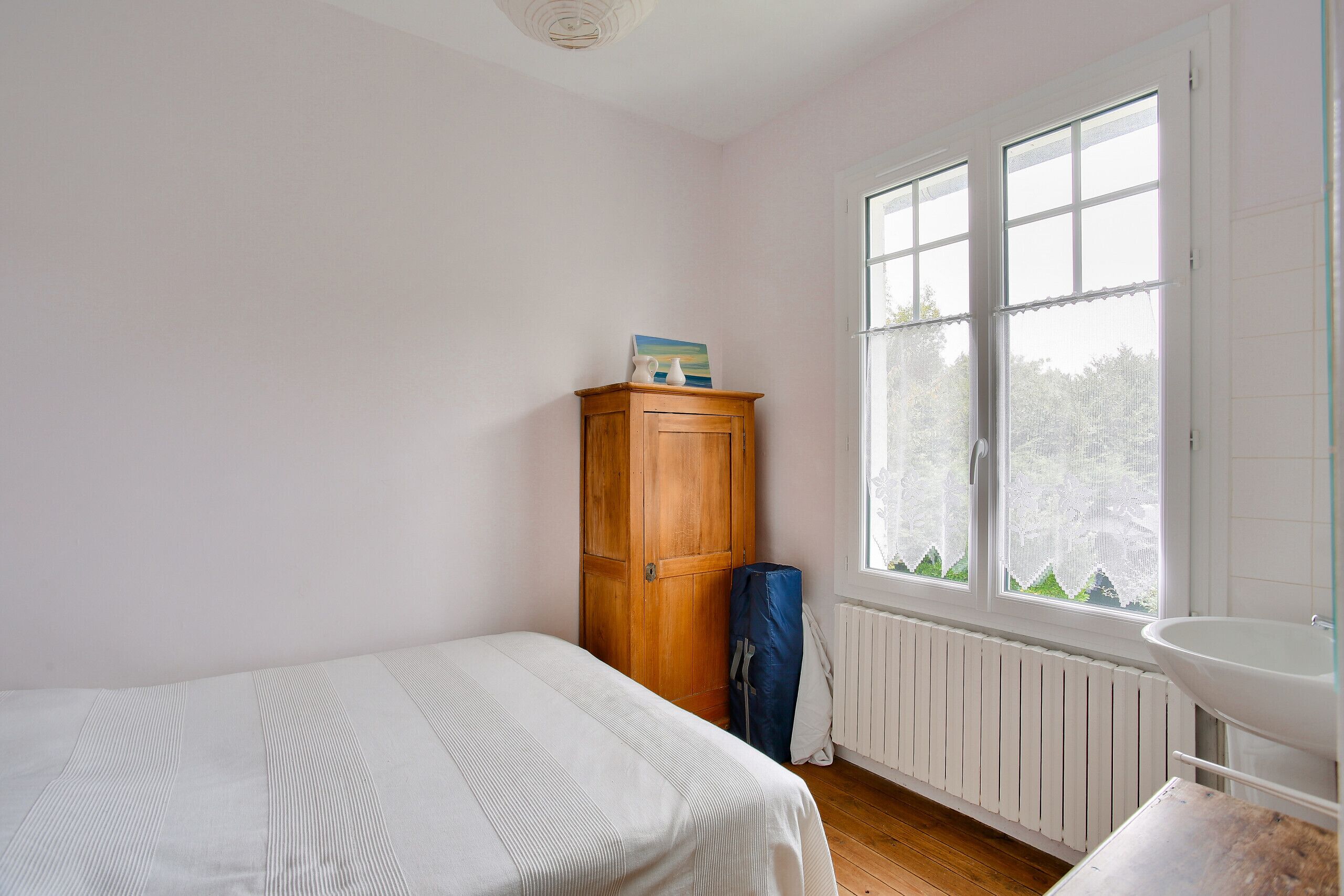 Een slaapkamer met een bed, een houten garderobe, een radiator en een raam met uitzicht op bomen.