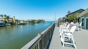 A waterfront promenade with a wooden deck, white Adirondack chairs, and a view of the water and buildings in the background. - Private Pool, Gas Grill, 2 Smokers, Boat Slip, Huge Deck & Patio & More! (Jamaica Beach)