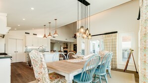 A spacious dining area with a wooden table and blue chairs, a kitchen in the background, and pendant lighting. - Private Pool, Gas Grill, 2 Smokers, Boat Slip, Huge Deck & Patio & More! (Jamaica Beach)