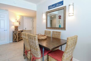 A dining area with a wooden table and wicker chairs, a wall-mounted mirror, and a sign reading 'Beach House.' - Incredible 10th Floor Balcony Views at Destin's Silver Shells! (Destin)