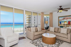 Ein Wohnzimmer mit Sofa, Sessel und Couchtisch, einem Deckenventilator und einem großen Fenster mit Blick auf das Meer und Gebäude.