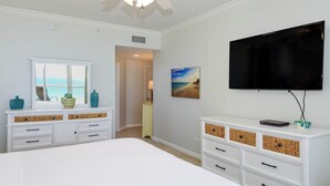 Un dormitorio con cama, cómoda blanca, televisor y un cuadro de una playa.