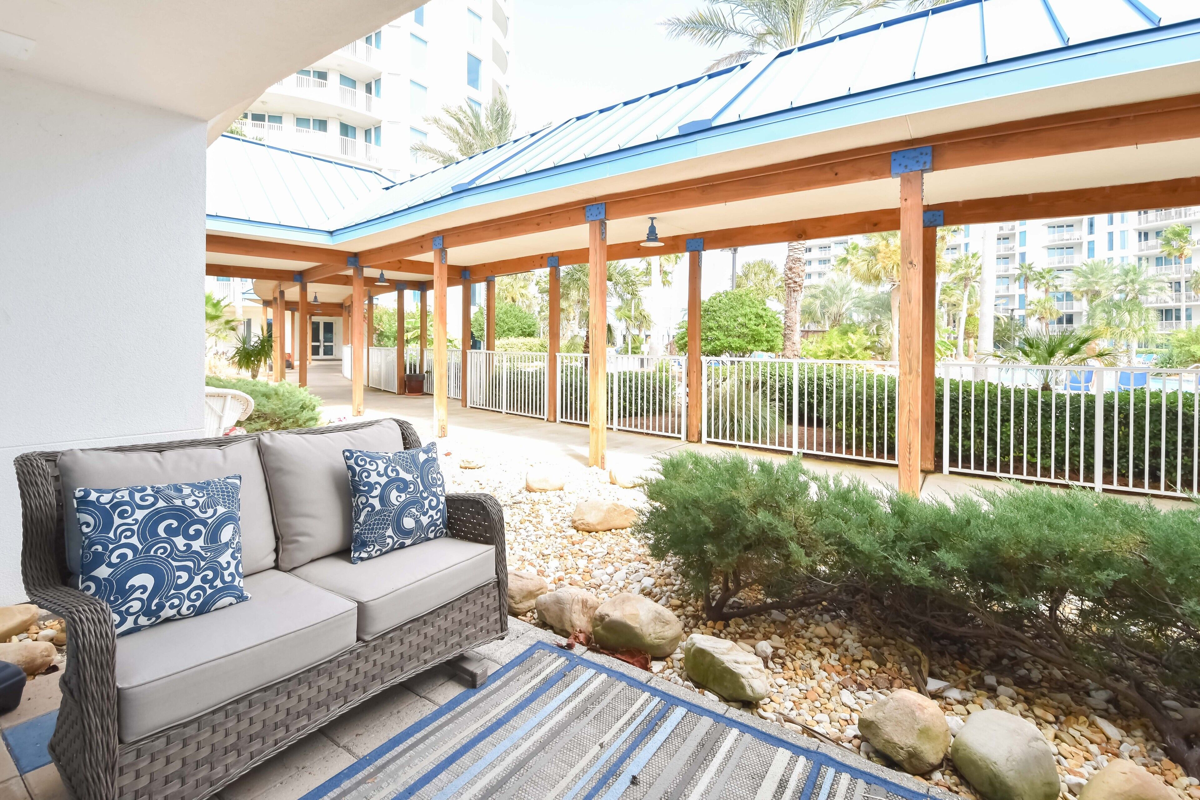 A covered outdoor seating area with a sofa and patterned cushions, surrounded by a metal fence and small plants.