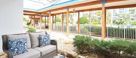 A covered outdoor seating area with a sofa and patterned cushions, surrounded by a metal fence and small plants.
