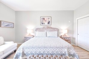 A bedroom with a large bed, two bedside tables with lamps, a white chair, and a door.