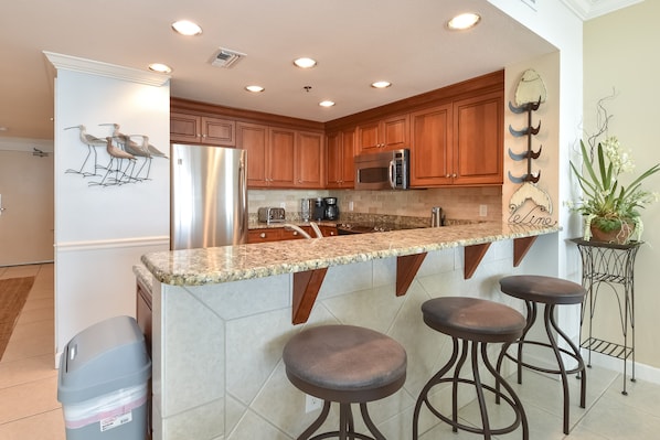 A kitchen with wooden cabinets, a granite countertop, a stainless steel refrigerator, a microwave, and a coffee maker. There are three bar stools and a potted plant.