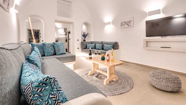 A modern living room with a grey sectional sofa, a wooden coffee table, and a flat-screen TV mounted on the wall. - Tiara Suites (Santorini)