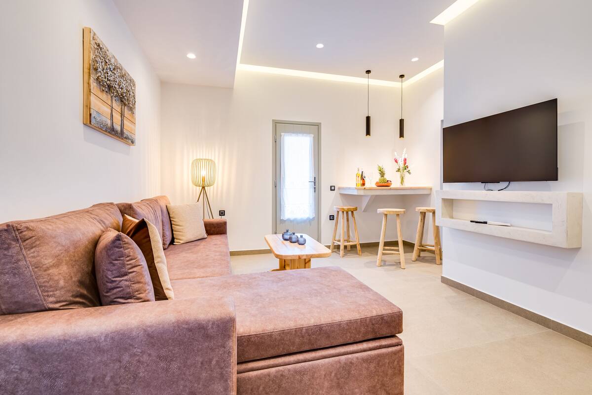 A modern living room with a sofa, a flat-screen TV mounted on the wall, and a dining area with stools and a table.