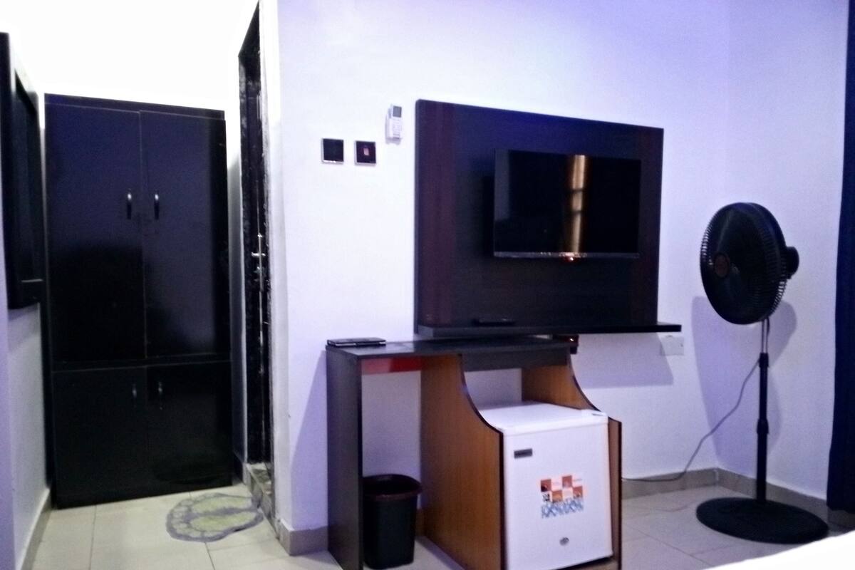 A hotel room with a bed, a desk with a computer, a TV mounted on the wall, a standing fan, and a wardrobe.